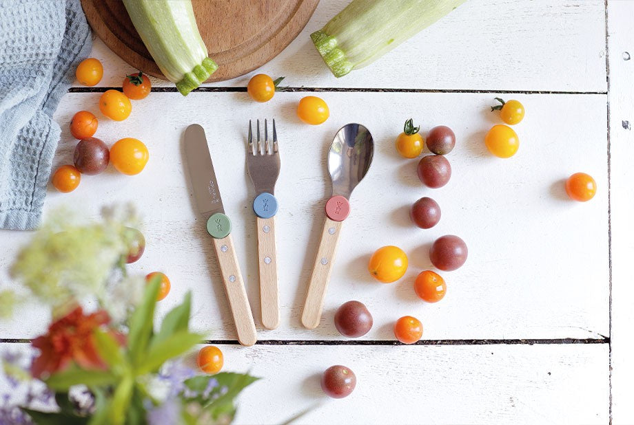 Couteau, fourchette et cuillère pour enfants Opinel disposés à plat sur une table avec des tomates cerises.