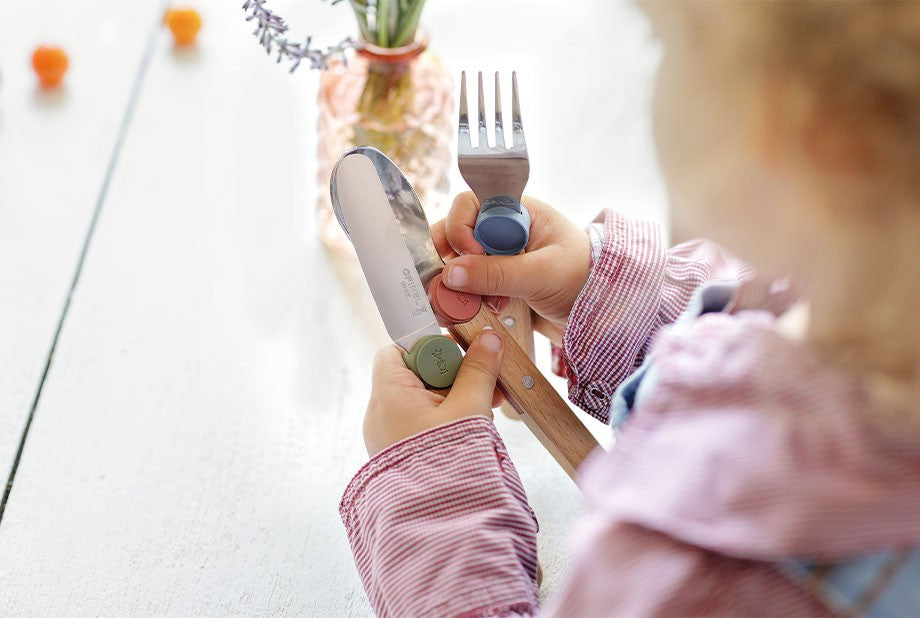 Enfant en bas âge tenant en main le couteau et la fourchette Opinel pour apprendre à manger de façon autonome.