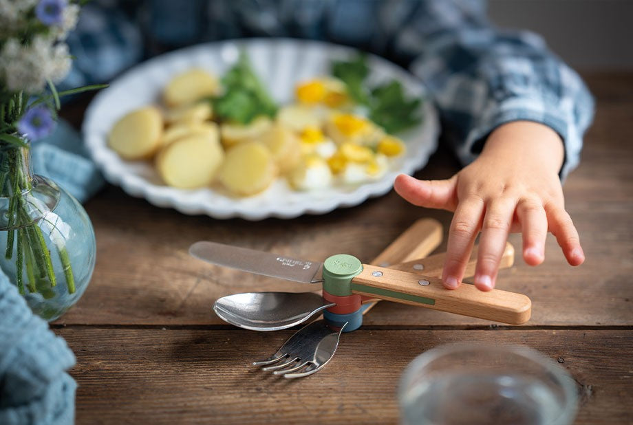Main d'enfant s'approchant du set de couverts Opinel Le Petit Gourmet assemblé sur une table en bois.